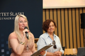 Dwie kobiety stoją na scenie, jedna z nich przemawia do mikrofonu i trzyma dokumenty w dłoni. W tle widoczne są elementy sali oraz baner Uniwersytetu Śląskiego.