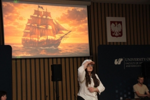 Na scenie stoi młoda dziewczyna, a w tle wyświetlony jest obraz żaglowca na morzu. Obok znajdują się banery Uniwersytetu Śląskiego oraz stanowisko muzyczne.