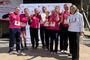Grupa dziewcząt w jednakowych różowych koszulkach trzyma puchar, dyplom i medale. Zdjęcie wykonano przed tablicami oraz ścianką z logotypami organizatorów.