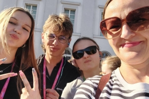 Cztery osoby pozują do selfie przed jasnym budynkiem z wysokimi oknami i ozdobną elewacją. 