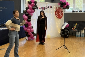 Dwie osoby stoją w sali przed dekoracją z czarnych i różowych balonów oraz białym tłem z napisem i rysunkiem serca. Jedna osoba ma na sobie ciemny strój z peleryną.