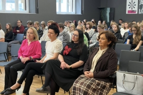 Kilka osób siedzi w pierwszym rzędzie sali konferencyjnej, a za nimi widoczna jest publiczność. Uczestnicy siedzą na krzesłach ustawionych w rzędach.