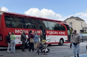 Czerwony autobus mobilnego punktu poboru krwi stoi na parkingu. Obok pojazdu znajduje się kilka osób.