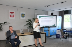 Kobieta stoi z mikrofonem przed ekranem z prezentacją w sali konferencyjnej. Obok siedzi tłumacz języka migowego.W tle widoczne są godła, rośliny i stół.