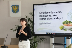 Kobieta stoi z mikrofonem obok ekranu z prezentacją o zdrowym żywieniu. W tle widoczne są rośliny oraz herb.
