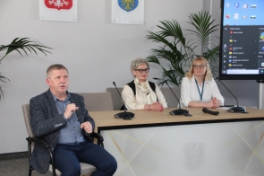 Trzy osoby siedzą przy stole konferencyjnym z mikrofonami. Po lewej stronie tłumacz języka migowego wykonuje gesty podczas wypowiedzi.
