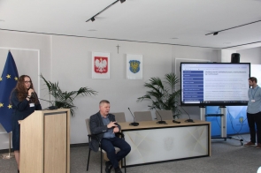 Kobieta przemawia przy mównicy w sali konferencyjnej. Obok siedzi tłumacz języka migowego, a po prawej stronie widoczny jest ekran z prezentacją i osoba stojąca przy monitorze.