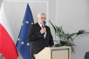Mężczyzna w garniturze stoi przy mównicy i przemawia do mikrofonu. W tle widoczne są flagi.