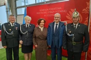 Pięć osób stoi przed czerwonym tłem z napisem Komendy Powiatowej Państwowej Straży Pożarnej w Cieszynie.