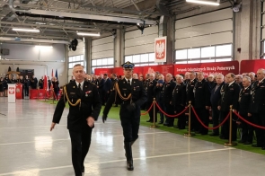 Dwóch strażaków w galowych mundurach maszeruje w hali podczas uroczystości. Po prawej stronie stoją uczestnicy wydarzenia, oddzieleni czerwonymi barierkami.