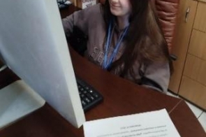Młoda dziewczyna siedzi przy biurku i pracuje przy komputerze stacjonarnym. Ma na sobie ciemną bluzę i identyfikator na niebieskiej smyczy. Na biurku leży kartka z informacjami o stażu uczniowskim, a w tle widać szafki oraz pudełka z dokumentami.
