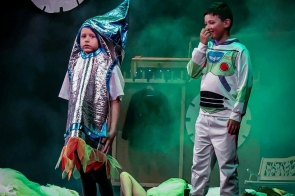 Na scenie stoi dwoje dzieci w kostiumach. Jedno dziecko ma srebrny strój rakiety z kolorowymi elementami przy nogach, a drugie ubranie przypominające strój astronauty. W tle, na podłodze sceny, leżą osoby w jasnych strojach.