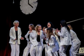 Na scenie stoi grupa kobiet ubranych w białe garnitury i czarne bluzki. Kobiety stoją blisko siebie, gestykulują, część z nich się śmieje.