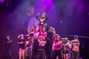 Na scenie stoi grupa młodych osób ustawionych w choreograficznej pozie. W centrum, na podwyższeniu, znajduje się dziewczyna w czarnym stroju i różowym topie, unosząca ręce do góry. Pozostałe osoby stoją wokół niej w czarnych i różowych ubraniach, wykonując podobne gesty rękami.