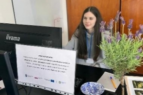 Młoda dziewczyna siedzi przy biurku w pomieszczeniu biurowym i pracuje przy komputerze. Na biurku znajdują się dwa monitory, notatnik, długopis oraz doniczka z lawendą. W tle widać drewniane szafki.
