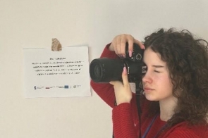 Młoda dziewczyna z kręconymi włosami trzyma aparat fotograficzny przy twarzy i wykonuje zdjęcie. Ubrana jest w czerwony sweter i ma identyfikator na szyi. Obok niej na ścianie znajduje się kartka z informacjami i logotypami projektu.