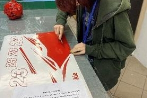 Młoda dziewczyna siedzi przy stole roboczym i przycina czerwono-białą folię reklamową za pomocą nożyka. Na stole leżą przygotowane elementy z napisami oraz kartka z informacjami o projekcie.