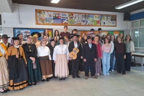 Duża grupa osób pozuje do wspólnego zdjęcia w sali. Część uczestników ubrana jest w tradycyjne stroje, a jedna osoba trzyma gitarę. W tle widać kolorową gazetkę ścienną, okna oraz szkolne wnętrze.
