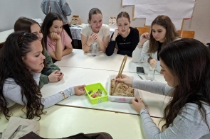 Kilka osób siedzi przy stole podczas warsztatów. Uczestniczki obserwują doświadczenie lub zadanie wykonywane w pojemniku z piaskiem, używając drewnianych szczypiec. Na stole znajdują się także kolorowe przedmioty, butelki z wodą i telefony.