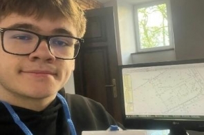Młody mężczyzna w okularach siedzi przy biurku i trzyma identyfikator stażysty zawieszony na niebieskiej smyczy. Obok znajduje się komputer z widocznym projektem lub planem technicznym na ekranie.
