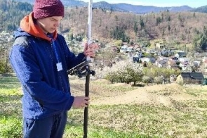 Młody mężczyzna stoi na wzgórzu i obsługuje urządzenie geodezyjne zamontowane na wysokim statywie. Ma na sobie czapkę, kurtkę oraz identyfikator zawieszony na szyi.