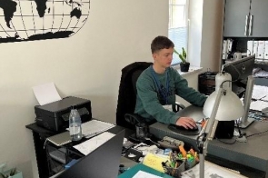 Młody mężczyzna siedzi przy biurku i pracuje przy komputerze w biurze. Na ścianie znajduje się grafika przedstawiająca mapę świata. W pomieszczeniu stoją biurka z monitorami, drukarka oraz lampki biurkowe.