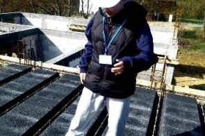 Młoda osoba w białym kasku ochronnym stoi na placu budowy na betonowej konstrukcji z widocznymi zbrojeniami i elementami izolacyjnymi. Ma na sobie ciemną kurtkę, jasne spodnie oraz identyfikator zawieszony na szyi.