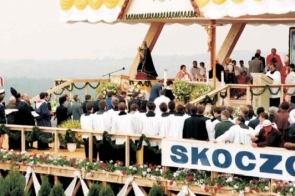 Na pierwszym planie widoczny jest tłum osób stojących przed podwyższeniem ozdobionym kwiatami i napisem „SKOCZÓW”.