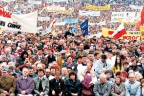 Tłum ludzi zgromadzony na otwartym terenie podczas dużego wydarzenia. Nad uczestnikami widoczne są liczne transparenty, flagi i banery.