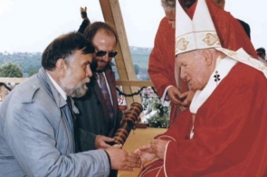 Jan Paweł II w czerwonych szatach siedzi na podwyższeniu i podaje rękę mężczyźnie. W tle stoją inne osoby uczestniczące w uroczystości.