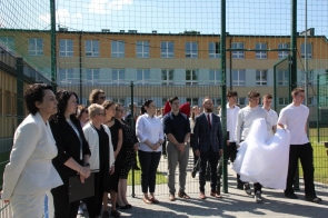 Grupa osób stoi przy wejściu na boisko sportowe. Po prawej stronie uczniowie trzymają biało-czerwoną tkaninę, a w tle widoczne jest ogrodzenie oraz budynek szkoły.