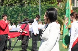 Kornelia Zubek Dyrektor ZSGH w Wiśle stoi przy mikrofonie na boisku sportowym. W tle widoczni są uczniowie ustawieni w rzędach oraz osoby trzymające czerwony materiał.