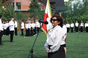 Janina Żagan Starosta Cieszyński stoi przy mikrofonie na boisku sportowym. Za nią widoczny jest poczet sztandarowy oraz uczniowie ustawieni w rzędach na murawie.