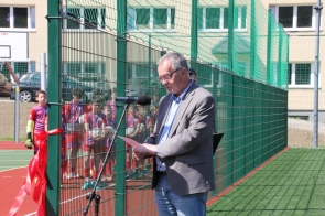 Aleksy Swoboda Skarbnik Powiatu stoi przy mikrofonie na boisku sportowym i trzyma kartkę w dłoniach. W tle, za ogrodzeniem boiska, widoczni są młodzi piłkarze w czerwono-niebieskich strojach.