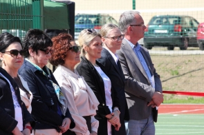 Przedstawiciele powiatu stoją obok siebie przy boisku sportowym. W tle widoczne są ogrodzenie, samochody oraz czerwona wstęga.