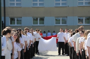 Uczniowie ubrani na biało stoją w dwóch rzędach na szkolnym dziedzińcu. Na środku kilka osób trzyma biało-czerwoną tkaninę, a w tle widoczny jest budynek szkoły.