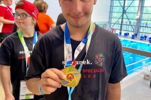 Młody mężczyzna stoi przy basenie i trzyma w dłoni złoty medal zawieszony na biało-niebieskiej wstążce. Ma na sobie czarną koszulkę z napisem „Olimpiady Specjalne Polska”.