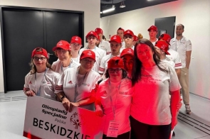 Grupa uczestników w białych koszulkach i czerwonych czapkach stoi w korytarzu hali sportowej. Z przodu kilka osób trzyma tablicę z napisem „Olimpiady Specjalne Polska BESKIDZKIE”.