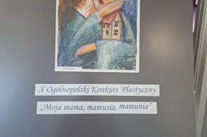 Na ciemnej planszy wystawowej widoczna jest praca plastyczna przedstawiająca kobietę trzymającą dom. Nad obrazem znajduje się napis „GRAND PRIX”. Poniżej umieszczono informacje o Konkursie Plastycznym. 