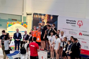 Grupa zawodników stoi na podium przy basenie i trzyma medale. Obok znajdują się trenerzy, organizatorzy oraz osoby robiące zdjęcia.