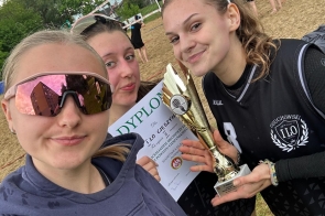 Trzy dziewczyny stoją na boisku do siatkówki plażowej. Dwie z nich trzymają puchar i dyplom, a w tle widoczne są siatka do gry oraz uczestnicy wydarzenia.
