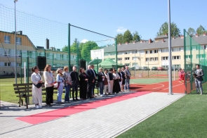 Grupa osób stoi przy boisku sportowym obok czerwonego dywanu. Po prawej stronie mężczyzna przemawia do mikrofonu, a w tle widoczne są budynki szkoły i ogrodzenie boiska.