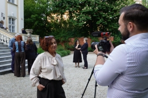 Janina Żagan Starosta Cieszyński stoi przed kamerą w ogrodzie przed pałacem. W tle widoczne są schody budynku oraz uczestnicy wydarzenia rozmawiający na dziedzińcu.