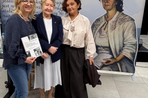 Trzy kobiety stoją przed banerem Centrum Edukacyjno-Kulturalnego „U Kossaków” w Górkach Wielkich. Jedna z osób trzyma książkę, a obok widoczna jest ilustracja kobiety.