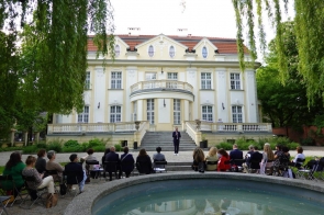 Widok na jasny pałac otoczony drzewami. Przed budynkiem ustawiono krzesła dla uczestników wydarzenia, a na schodach stoi przemawiająca osoba. Na pierwszym planie widoczna jest fontanna.