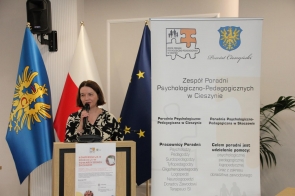Kobieta przemawia przy mównicy, trzymając mikrofon. Obok stoi baner Zespołu Poradni Psychologiczno-Pedagogicznych w Cieszynie z herbem Powiatu Cieszyńskiego i informacjami o działalności poradni. W tle widoczne są flagi.