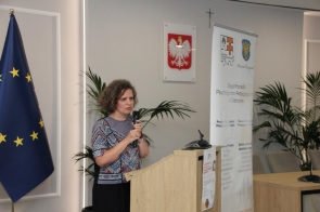 Kobieta stoi przy mównicy i przemawia do mikrofonu. Obok widoczny jest baner Zespołu Poradni Psychologiczno-Pedagogicznych w Cieszynie, a w tle flaga Unii Europejskiej, godło Polski i rośliny doniczkowe.