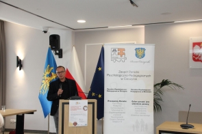 Mężczyzna w ciemnej marynarce stoi przy mównicy i przemawia do mikrofonu. Obok znajduje się baner, a w tle widoczne są flagi.