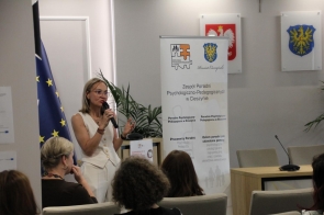 Kobieta w białym stroju przemawia do mikrofonu przed siedzącymi uczestnikami spotkania. Obok znajduje się baner Zespołu Poradni Psychologiczno-Pedagogicznych w Cieszynie.
