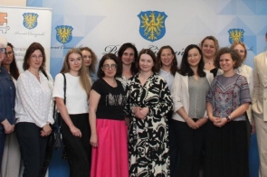 Grupa kobiet i mężczyzna w eleganckich strojach pozująca do zdjęcia na tle ścianki z banerów z herbem Powiatu Cieszyńskiego.